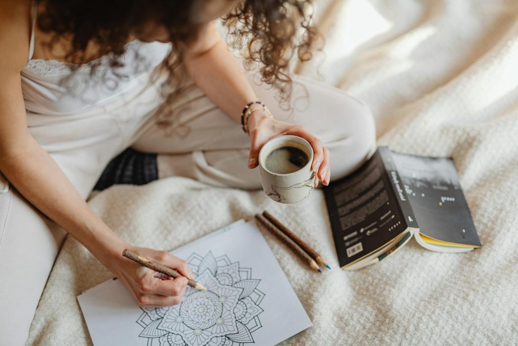 Woman coloring mandala while holding coffee on cozy blanket, enjoying a relaxing moment, coloring book with motivational quotes and colored pencils 